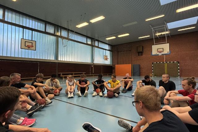Die Teilnehmenden sitzen in einem Kreis in einer Sporthalle, während die Basketballregeln erklärt werden.